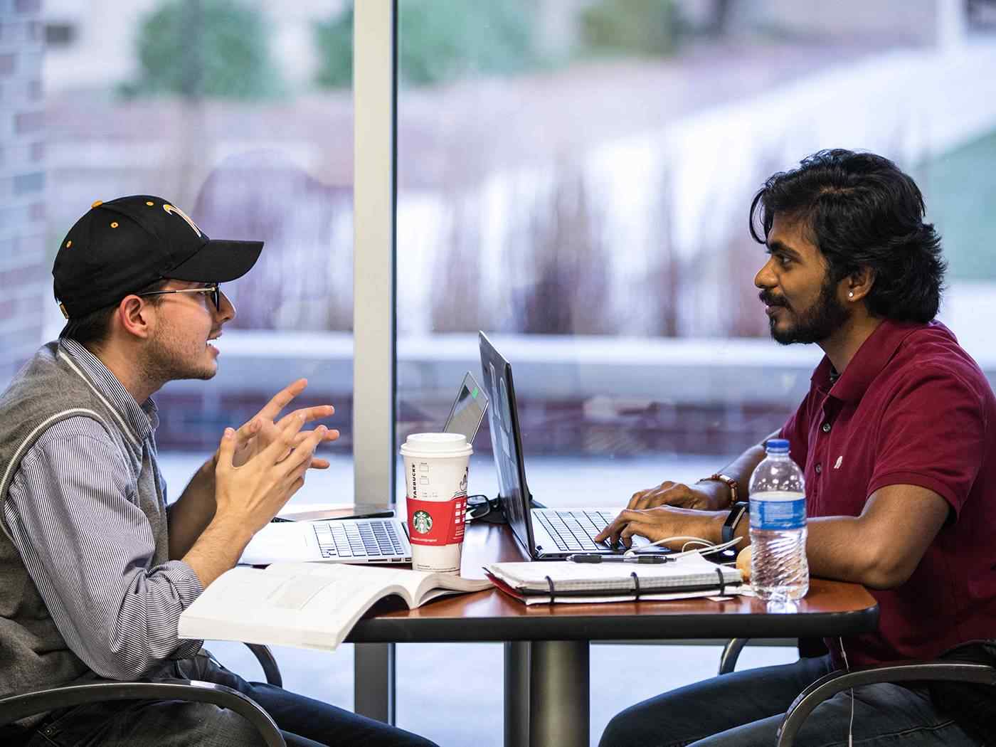 Two communications students at opposite ends of a table have have a lively conversation over their open laptop computers. 